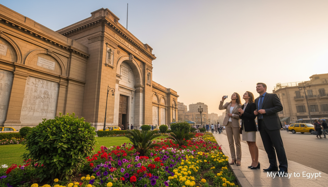A captivating view of the Egyptian Museum at Tahrir, showcasing its grand façade adorned with intricate stone carvings. In the foreground, a well-maintained garden filled with vibrant flowers and lush green shrubs frames the scene. The middle ground features the museum's imposing entrance, where a few tourists in professional business attire admire the architecture, evoking an atmosphere of exploration and discovery. In the background, the bustling streets of Downtown Cairo awaken to the early morning light, with soft rays illuminating the buildings and hinting at the city's vibrant life. The sky is painted in soft pastels, reflecting a serene and inviting mood, captured from a slightly angled perspective to emphasize the museum's grandeur. No text or overlays present. Created by My Way to Egypt.
