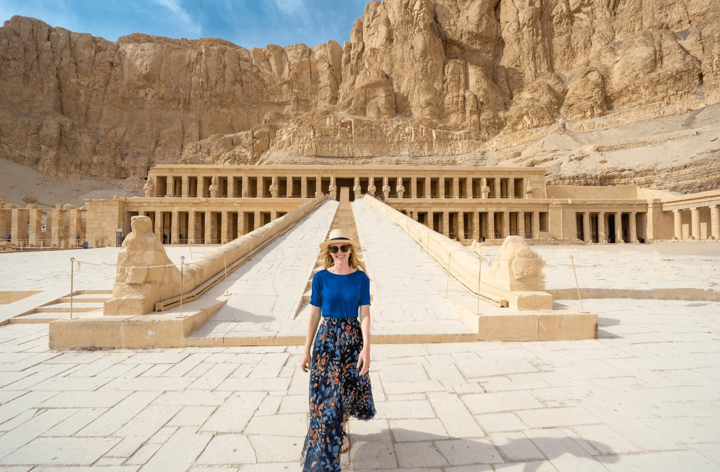 A smiling woman in a blue shirt and sun hat walking on the stone courtyard in front of the terraced Mortuary Temple of Hatshepsut in Luxor, Egypt.
