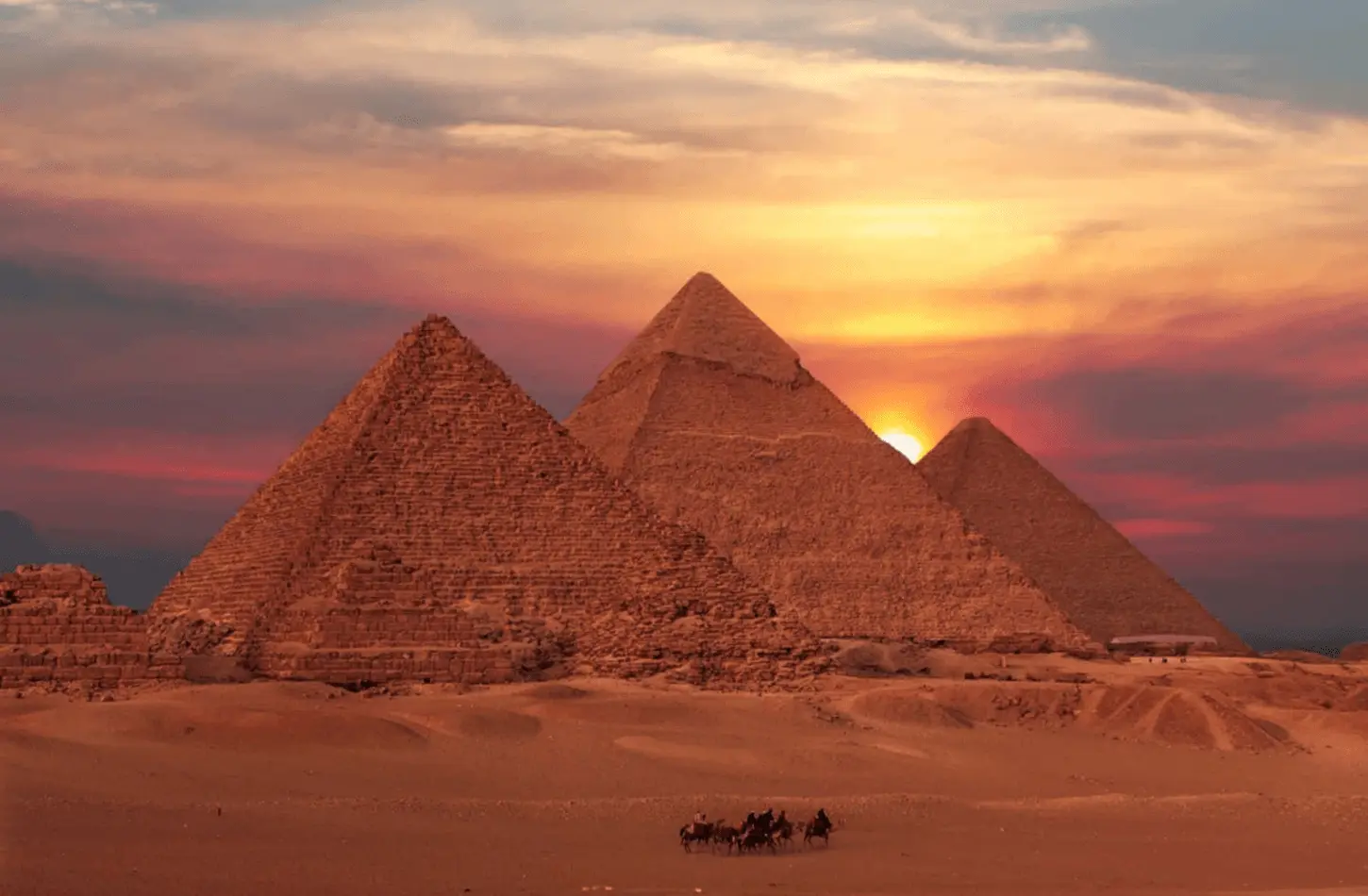 The three Great Pyramids of Giza at sunset with a camel caravan crossing the desert sand in the foreground.