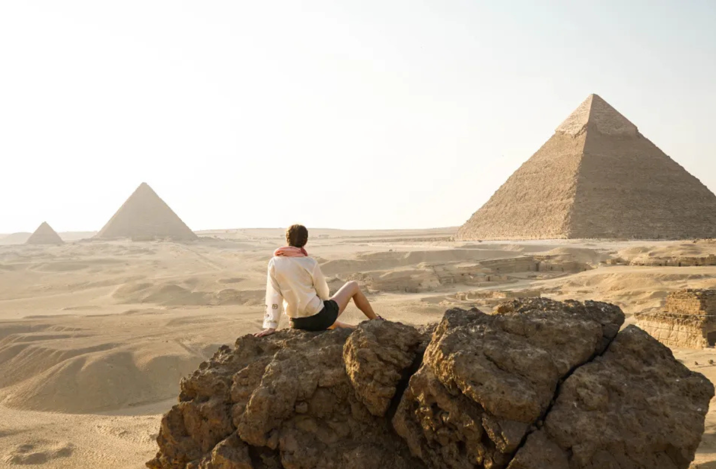 A traveler sitting on a rock overlooking the three Great Pyramids of Giza in the desert under a bright hazy sky