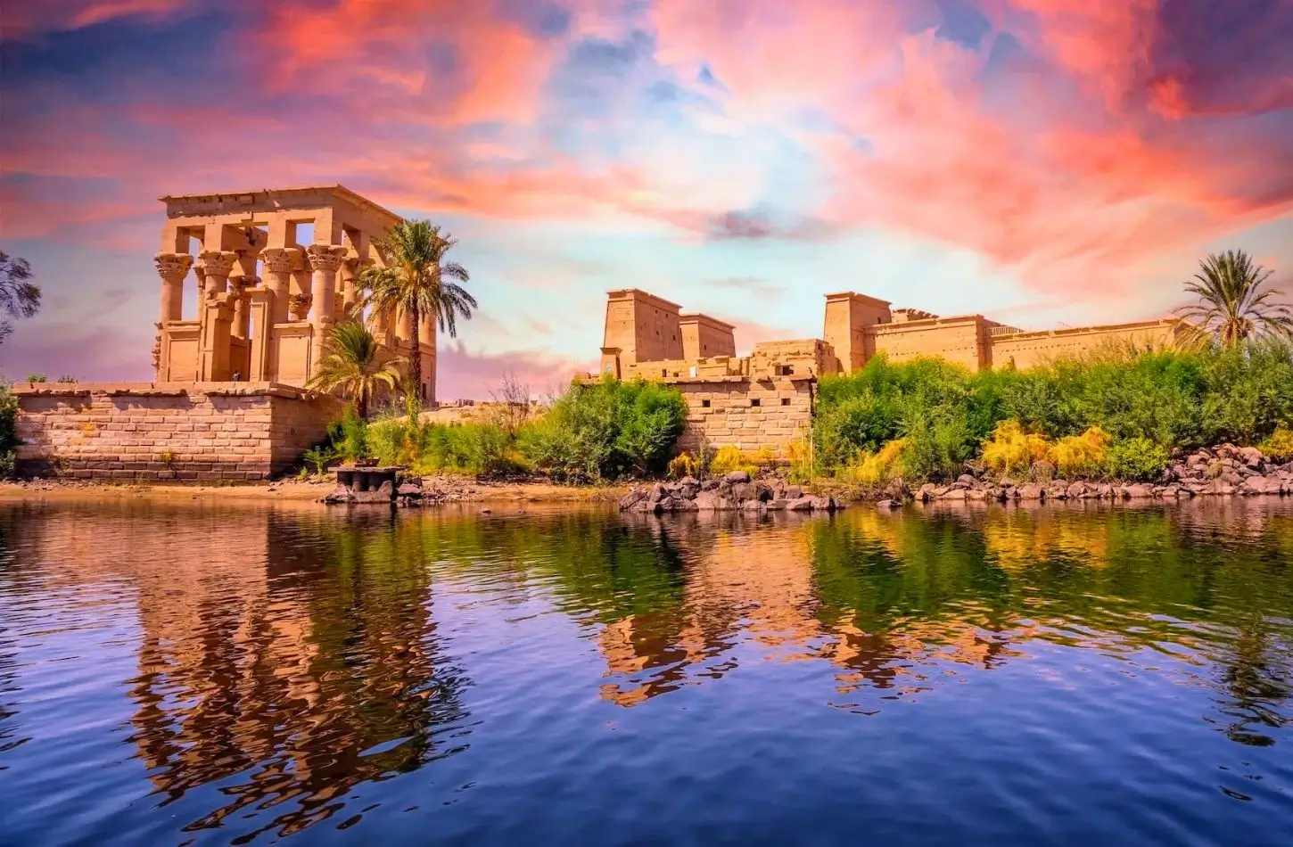 Philae Temple complex and Trajan's Kiosk reflecting on the Nile River during a vibrant sunset.