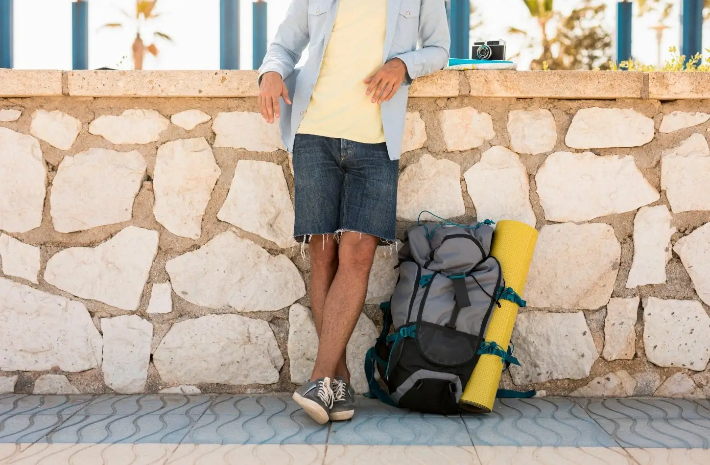 A solo backpacker in denim shorts and a light shirt leans against a stone wall next to a large travel backpack and yellow sleeping mat, representing gear from an Egypt packing list.