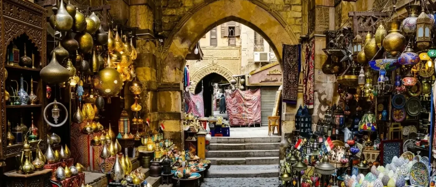 Vibrant market stalls filled with traditional brass lamps, colorful mosaic lanterns, and handcrafted plates under a stone archway at Khan El-Khalili bazaar, showcasing the best Egypt souvenirs.