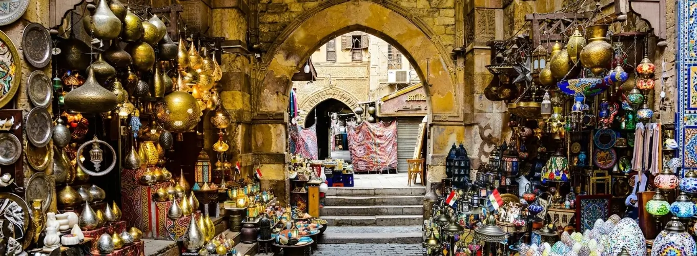 Vibrant market stalls filled with traditional brass lamps, colorful mosaic lanterns, and handcrafted plates under a stone archway at Khan El-Khalili bazaar, showcasing the best Egypt souvenirs.