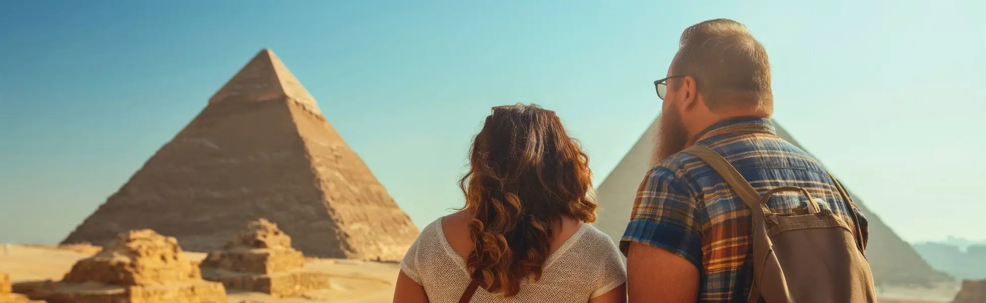 A couple from behind looks out at the Great Pyramids of Giza in the desert under a clear blue sky, dressed in lightweight travel clothes perfect for an Egypt packing list.