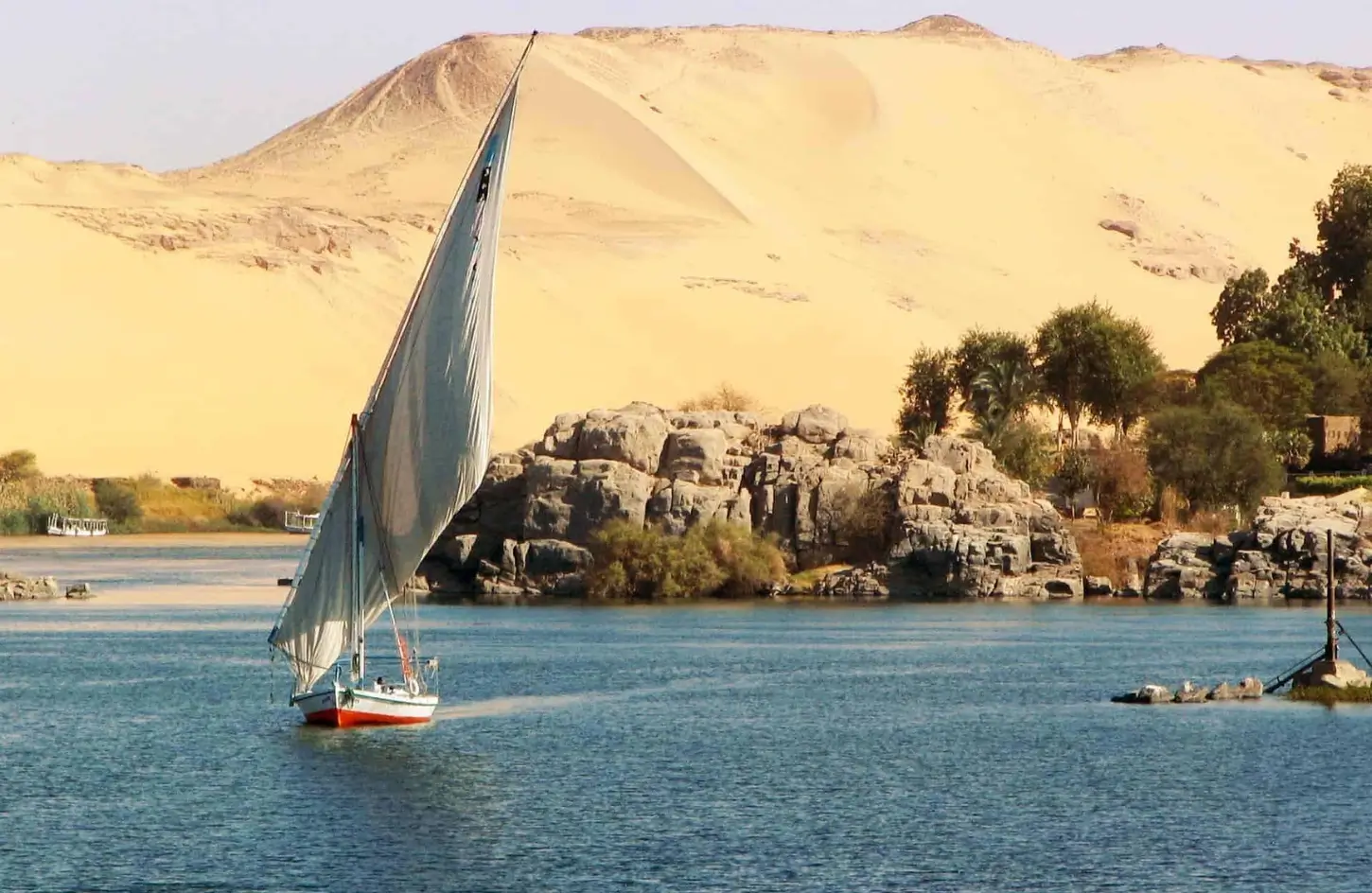 A traditional white-sailed felucca boat cruising down the Nile River in Aswan with golden desert sand dunes in the background, showing optimal weather conditions for visiting Egypt in October.