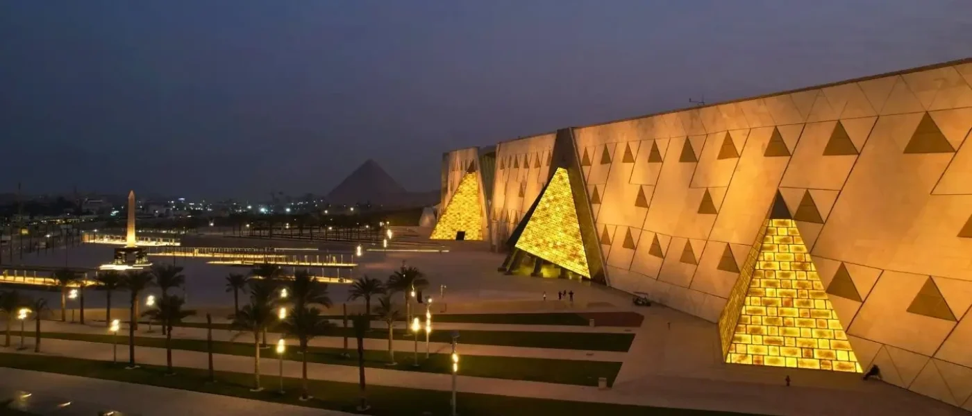 Night view of the modern Grand Egyptian Museum facade illuminated with warm golden lights, featuring translucent pyramid-shaped structures and the Giza pyramids visible in the background dusk.