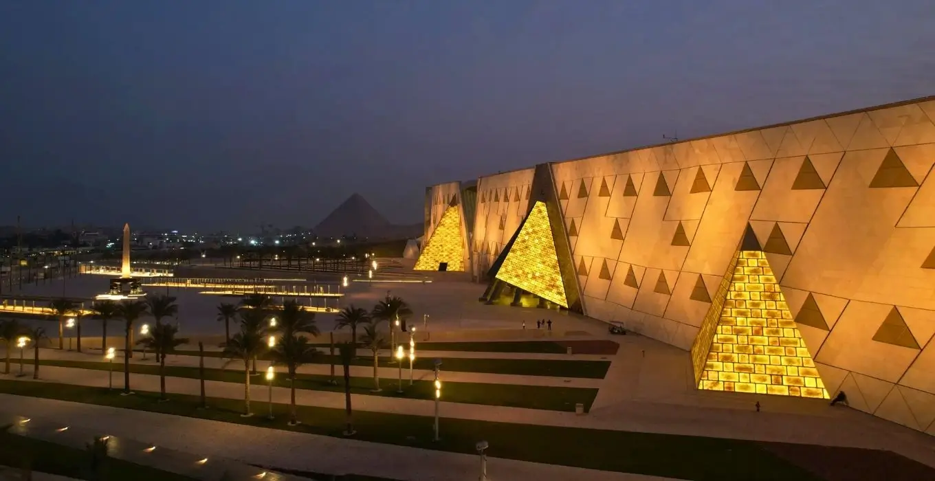 Night view of the modern Grand Egyptian Museum facade illuminated with warm golden lights, featuring translucent pyramid-shaped structures and the Giza pyramids visible in the background dusk.