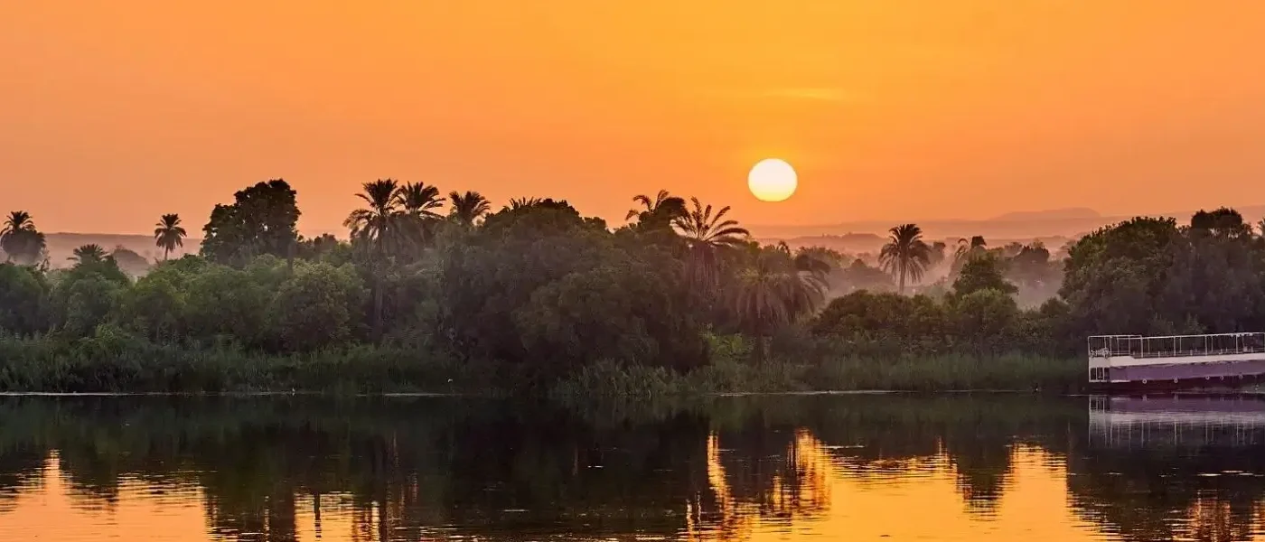 Golden hour sunset over the Nile River with palm trees and a boat, showcasing scenic routes for how to get around Egypt.