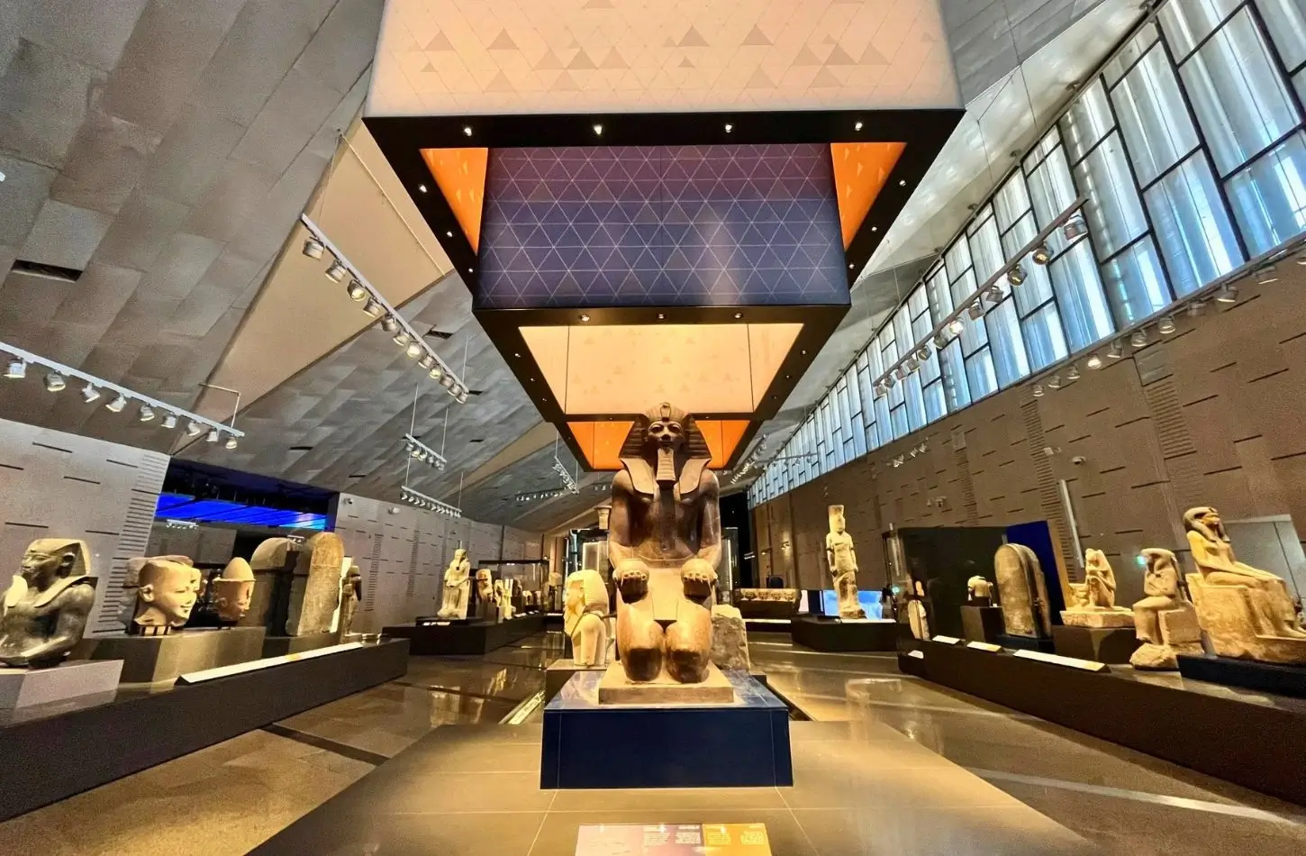 A central pharaoh statue under a large, illuminated geometric canopy inside a modern exhibition hall at the Grand Egyptian Museum, surrounded by ancient artifacts.