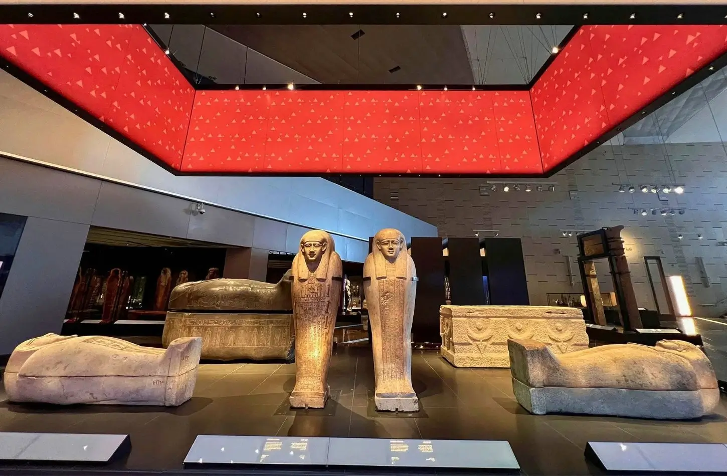 Ancient Egyptian stone sarcophagi and mummiform coffins on display inside the modern gallery of the Grand Egyptian Museum, beneath a large red geometric ceiling installation.