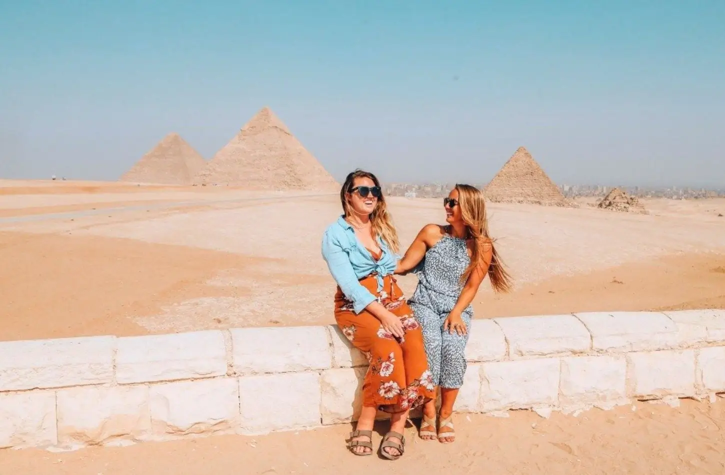 Two female travelers laugh while sitting on a stone wall with the Giza Pyramid complex in the background, wearing breathable outfits included in their Egypt packing list.