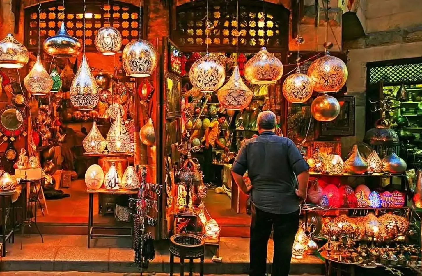 A shopkeeper stands in front of a vibrant stall filled with glowing, intricate brass lamps and fanous lanterns at Khan el-Khalili bazaar, showcasing one of the top things to do in Egypt.