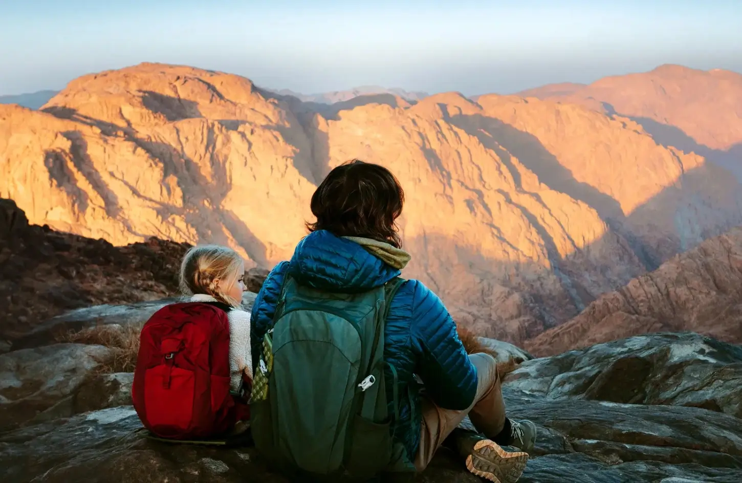Two hikers sitting on a rocky summit with backpacks, looking out over the rugged mountain peaks of Mount Sinai during sunrise in Egypt in September.