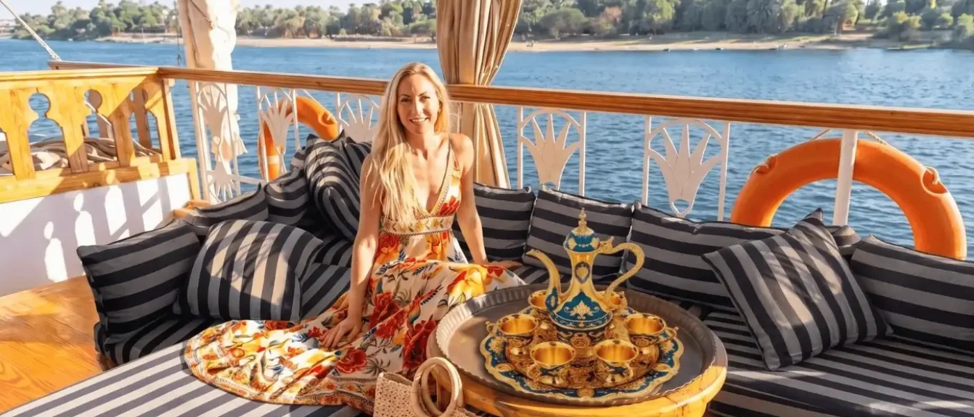 Dahabiya vs Nile Cruise: Woman relaxing on a wooden sailboat deck enjoying tea, illustrating the calm, intimate atmosphere of a Dahabiya sailing boat compared to a standard Nile cruise.