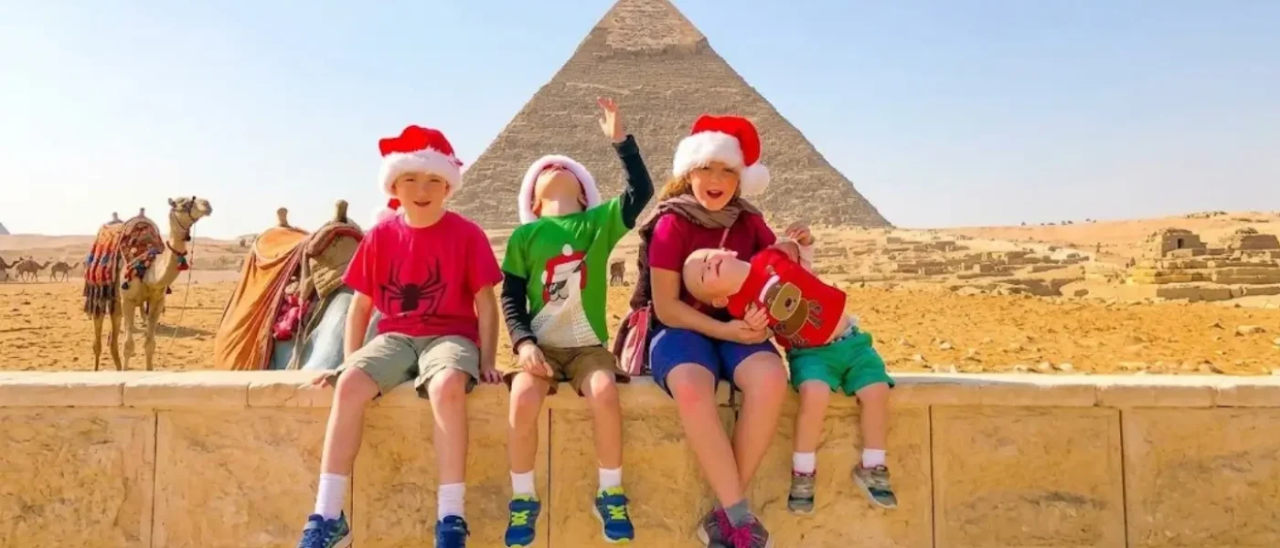 Children wearing Santa hats sitting in front of the Giza Pyramids while enjoying family holiday activities during Christmas in Egypt.