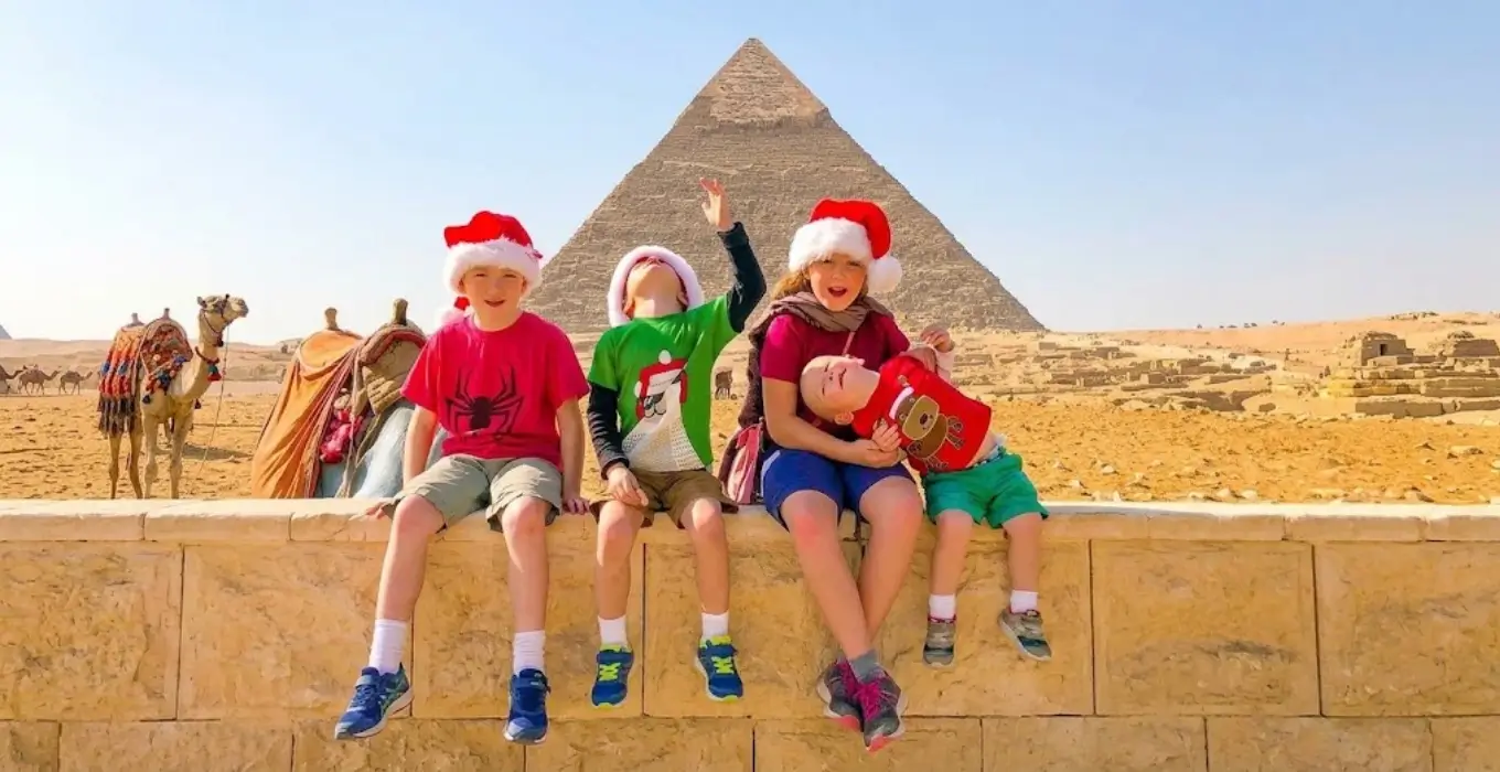 Children wearing Santa hats sitting in front of the Giza Pyramids while enjoying family holiday activities during Christmas in Egypt.