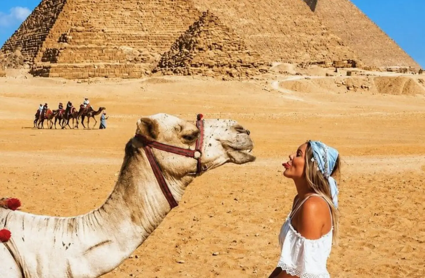 A woman in a white top and blue headband playfully stickout her tongue at a camel, in a close-up photo in front of the Great Pyramids during a visit to Egypt in February